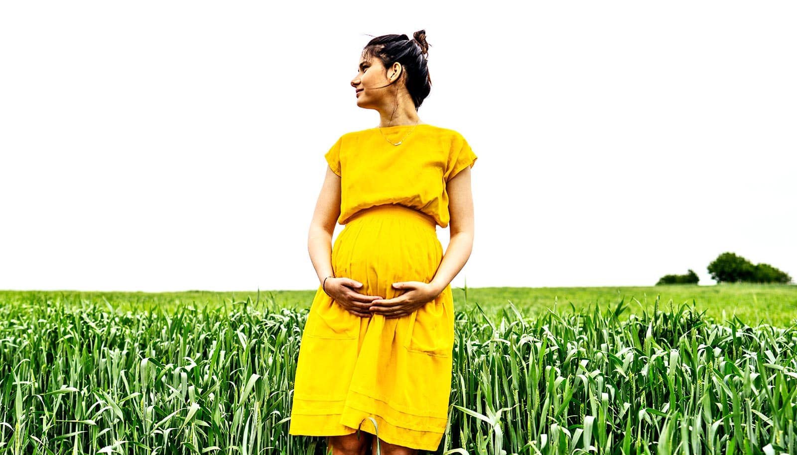 A pregnant woman in a yellow dress stands outside in a field of green grass