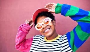 A woman wears a bright, colorful sweater and yellow-tinted sunglasses while smiling and touching her baseball cap.