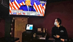 A woman wearing a surgical mask looks up at the TV above a cash register to see Donald Trump speaking in front of US flags.