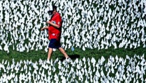 A man in red walks through a field of small white flags on green grass.