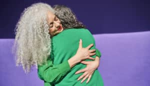 person with long silver hair hugs another person. Both wear green in front of purple background