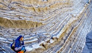 A researcher takes a picture while standing next to a cliff showing different levels of rock.