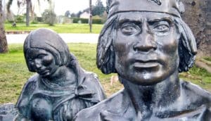 serious face of man with short hair and headband. Behind him, female figure holds infant