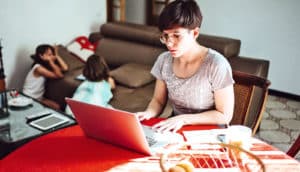 A woman works on a laptop sitting at her kitchen table, with kids playing on the floor in the background.