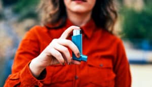 A woman in a dark orange button-up shirt holds a blue asthma inhaler.