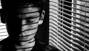 A teen boy has lines of shadow on his face from open blinds on the window next to him.