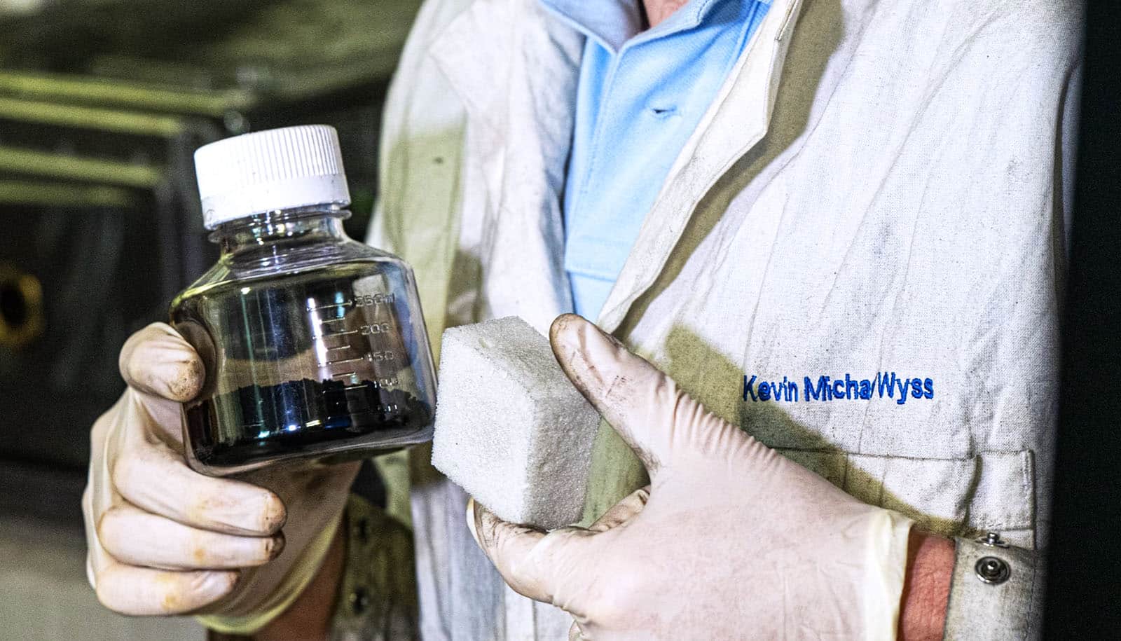 gloved hands hold flash of black substance and foam in front of lab coat