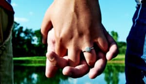A couple holds hands in front of a small lake with an engagement ring on one person's hand.