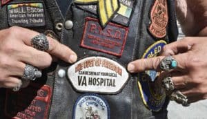 ringed hands point to patch on leather vest. Patch says "the cost of freedom can be seen at your local VA hospital"