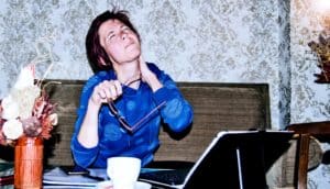 A woman takes off her glasses and holds her neck while wincing in pain.