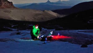 A researcher in winter gear crouches in the snow over equipment with green and red lights with mountains in the background.