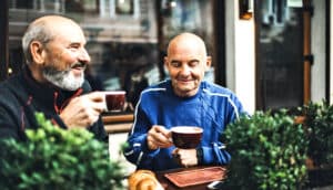 Two older men sit at an outdoor cafe table smiling and sipping coffee.
