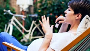 A pregnant woman blows on her nails and holds a bottle of nail polish while sitting outside.