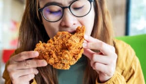 A young woman wearing glasses takes a bite out of a piece of fried chicken.