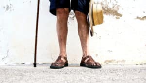 An older man stands against a wall holding a cane in one hand and a straw hat in the other with sandals on his feet.