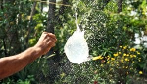 A person uses a pin to pop a water balloon hanging on a clothesline.