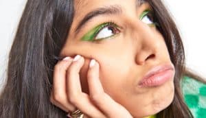 A woman wearing green eye makeup looks indecisive in front of a white background.