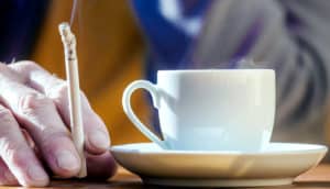 A person holds a cigarette next to a cup of coffee.