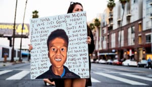 person holds sign with portrait of young Black man surrounded by his last words: "My name is Elijah McClain. I was just going home. I'm an introvert. I'm just different. That's all. I'm so sorry. I have no gun. I don't do that stuff. I don't do any fighting. Why are you attacking me? I don't even kill flies. I don't eat meat. But I don't judge people who do eat meat. Forgive me. All I was trying to do was become better. I will do it. I will do anything."