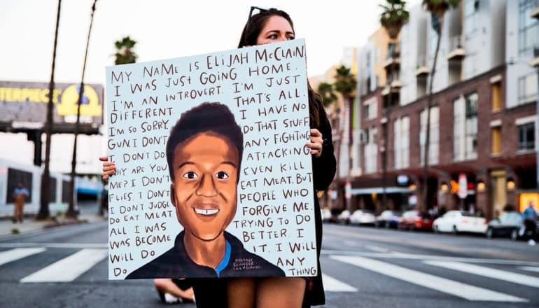 person holds sign with portrait of young Black man surrounded by his last words: "My name is Elijah McClain. I was just going home. I'm an introvert. I'm just different. That's all. I'm so sorry. I have no gun. I don't do that stuff. I don't do any fighting. Why are you attacking me? I don't even kill flies. I don't eat meat. But I don't judge people who do eat meat. Forgive me. All I was trying to do was become better. I will do it. I will do anything."