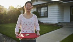 person smiles holding container of cookies in front of house
