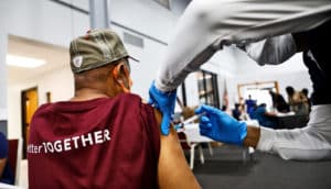 A man in a maroon t-shirt with the text "Better Together" on it gets a shot of the COVID-19 vaccine.