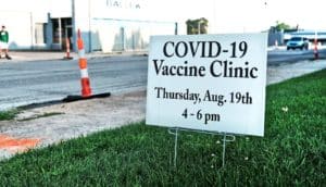 A sign for a COVID-19 vaccine clinic sits in some grass next to a vacant road.