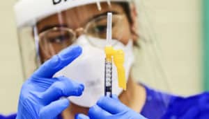 A health worker wearing a face shield, mask, and blue gloves flicks a syringe filled with a vaccine for monkeypox.