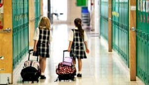 Two young girls wearing plaid school uniforms walk down a school hallway while rolling their backpacks.