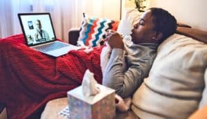 A man laying on a couch under a blanket holds a cup of tea while talking to a doctor on his laptop.