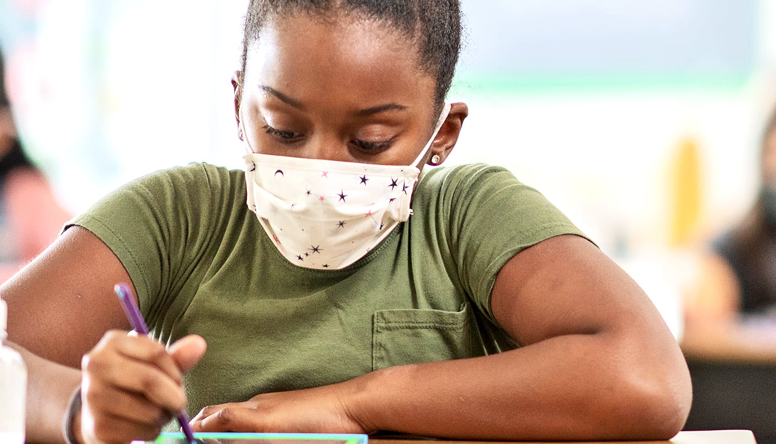 A young student wearing a face mask does schoolwork at a classroom desk.