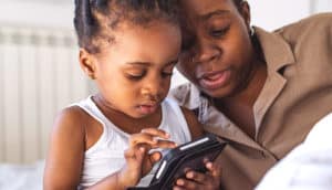 A mother and daughter look at a smart device while sitting on a bed.