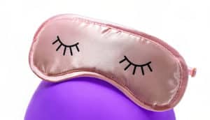 A pink sleep mask with two closed eyes printed on it sits on top of a purple balloon.