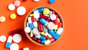 A bunch of different pills in a small bowl, spilling over onto an orange background.