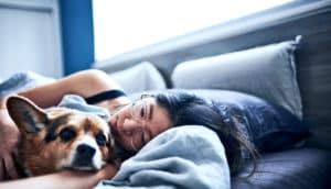 A young woman in bed with her dog while sunlight shines through the window above the bed.