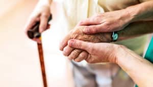 An older woman walking with a cane has another person holding her hand.