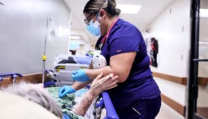 A nurse in scrubs stands beside a patient on a stretcher in a hospital hallway.