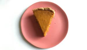 A slice of pumpkin pie sits on a pink plate against a white background.