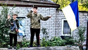 Two kids stand on a concrete barrier while waving to passing soldiers while holding the Ukrainian flag and a toy rifle.
