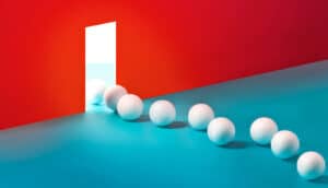 Ping pong balls in a line move through a small rectangular opening in a red wall.