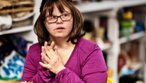 A woman wearing glasses and a purple top puts her hands together and looks off camera.