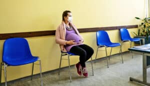 A pregnant woman sits in a doctor's office waiting room while wearing a medical face mask.