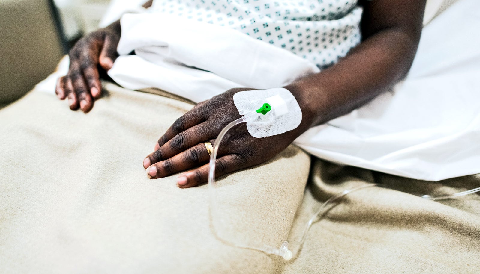 A Black patient in a hospital bed has a wedding ring and IV on the same hand.