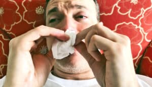 A man looks pained as he blows his nose with a tissue and lays on a red couch.