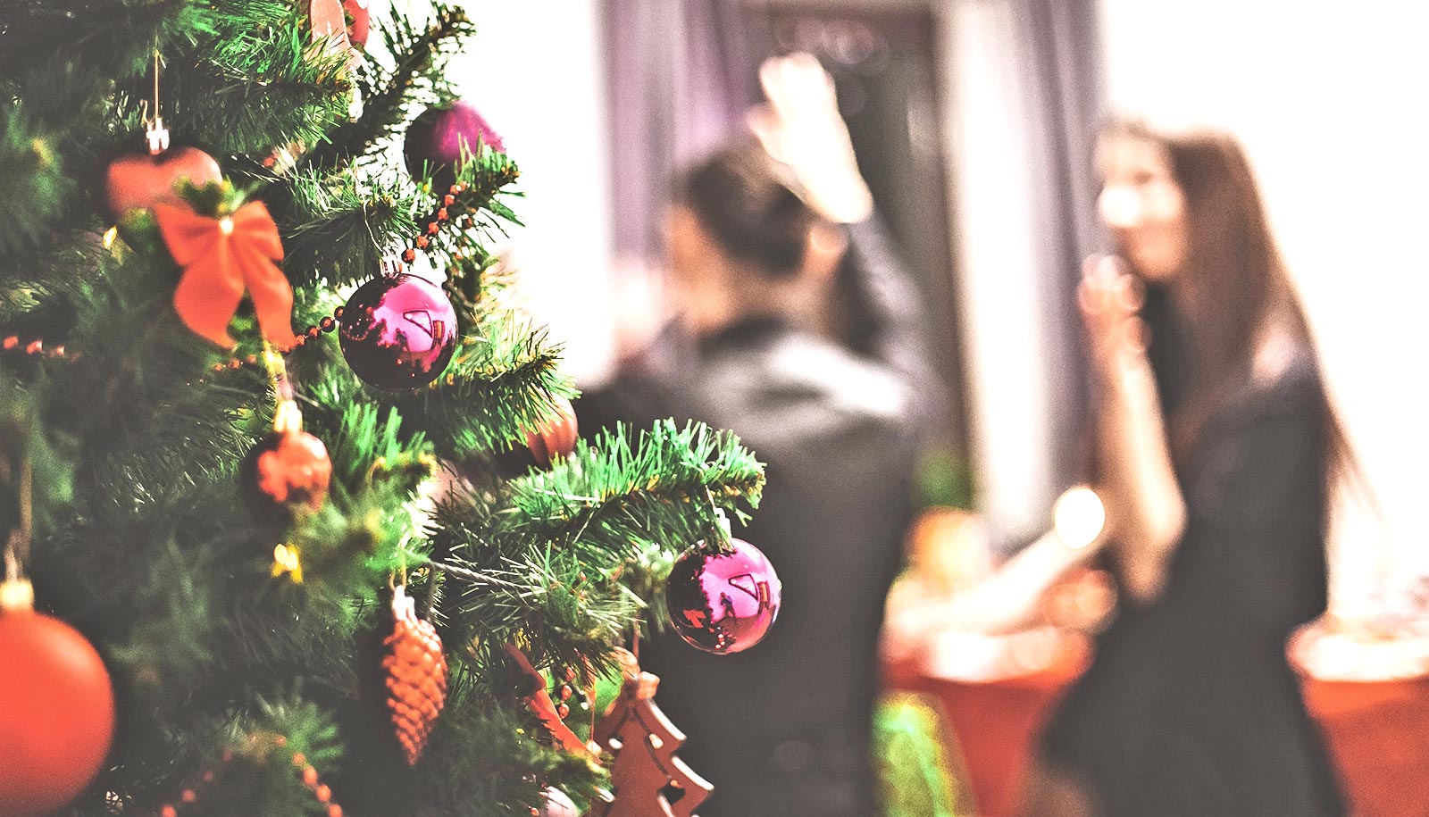 Et juletræ dækket af ornamenter med folk til en fest i baggrunden.