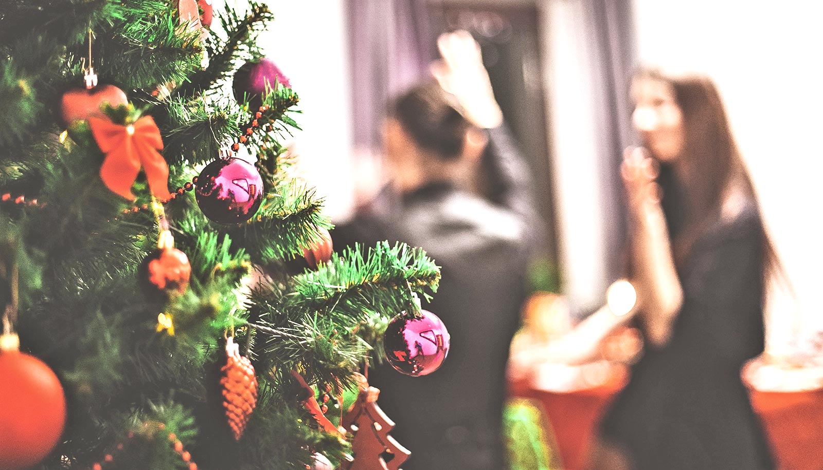 A Christmas tree covered in ornaments with people at a party in the background.