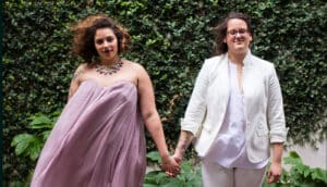 two brides in pale purple dress and white suit stand holding hands outdoors