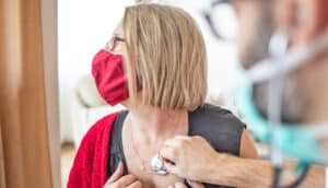 adult holds stethoscope to chest of middle-aged woman in red mask