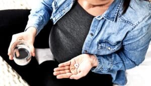 A pregnant woman holds a glass of water in one hand and white pills in the other.