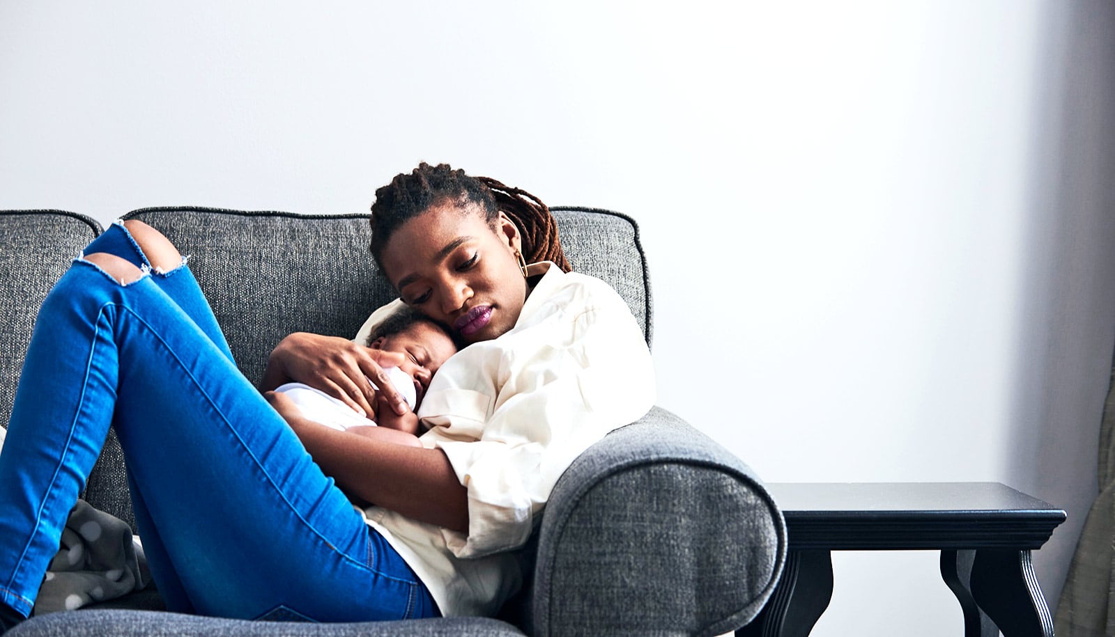 A woman holds her baby while sitting on a gray couch with her eyes closed.